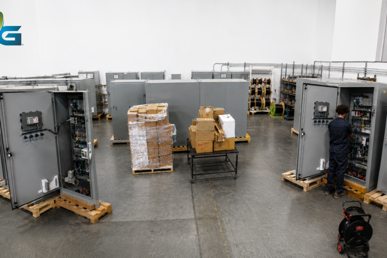 Warehouse with rows of open gray electrical control panels, a technician working at one panel and stacked pallets.