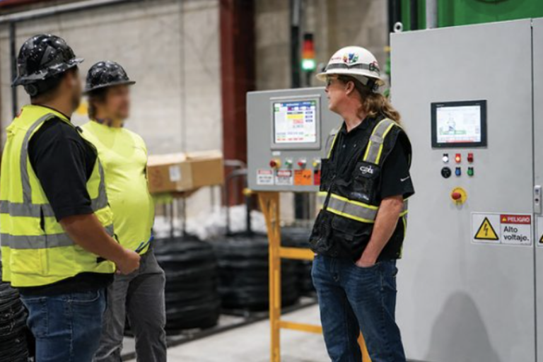 Three workers in hard hats and vests stand by control panels with touchscreens and high-voltage warning labels.