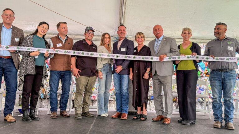 Eleven people stand side-by-side under a white tent holding a long ceremonial ribbon for a ribbon-cutting event.