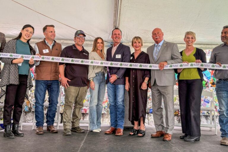 Eleven people stand side-by-side under a white tent holding a long ceremonial ribbon for a ribbon-cutting event.